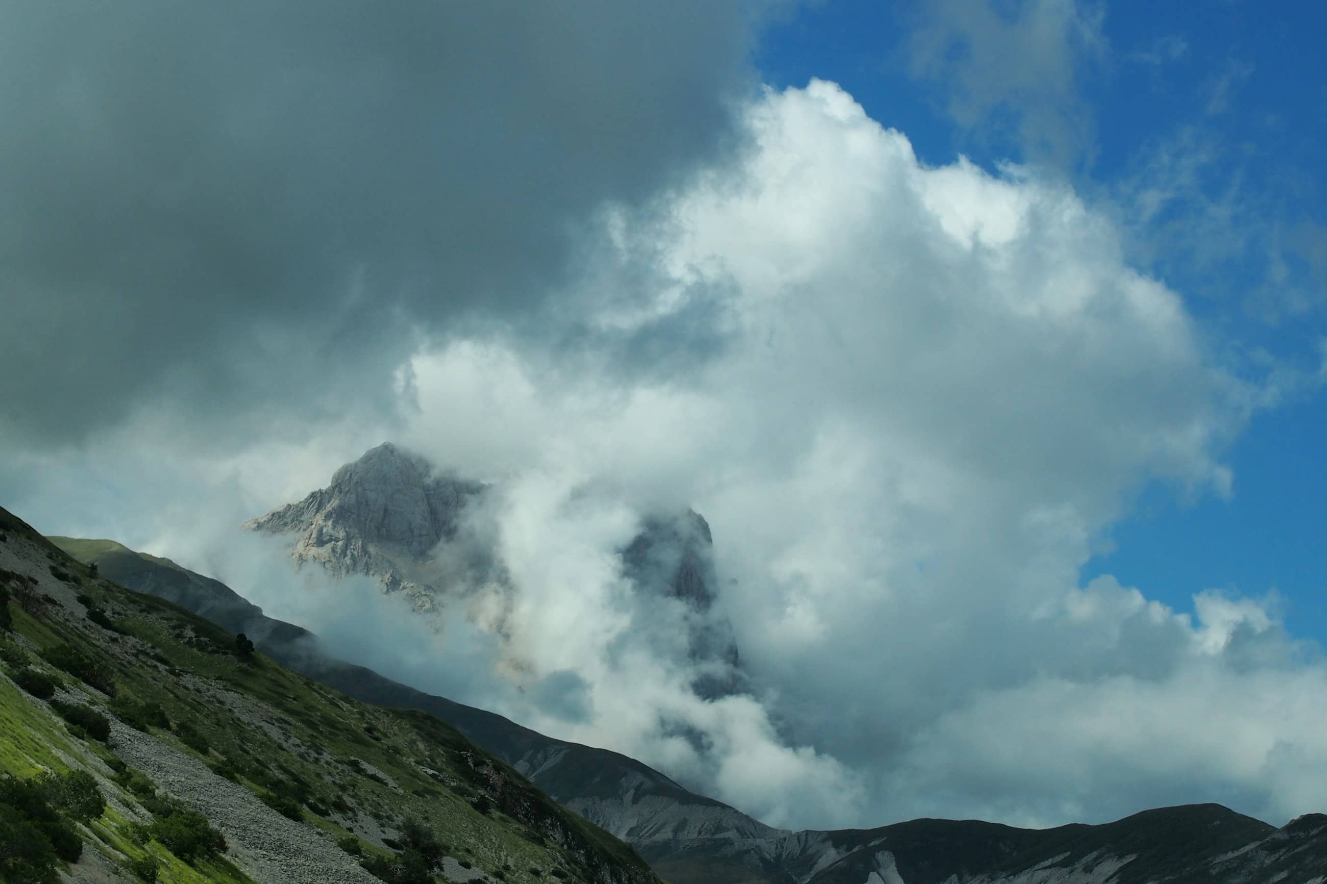 A grey rock summit of the Gran Sasso half-buried in cloud, dark forested slopes falling away below it.