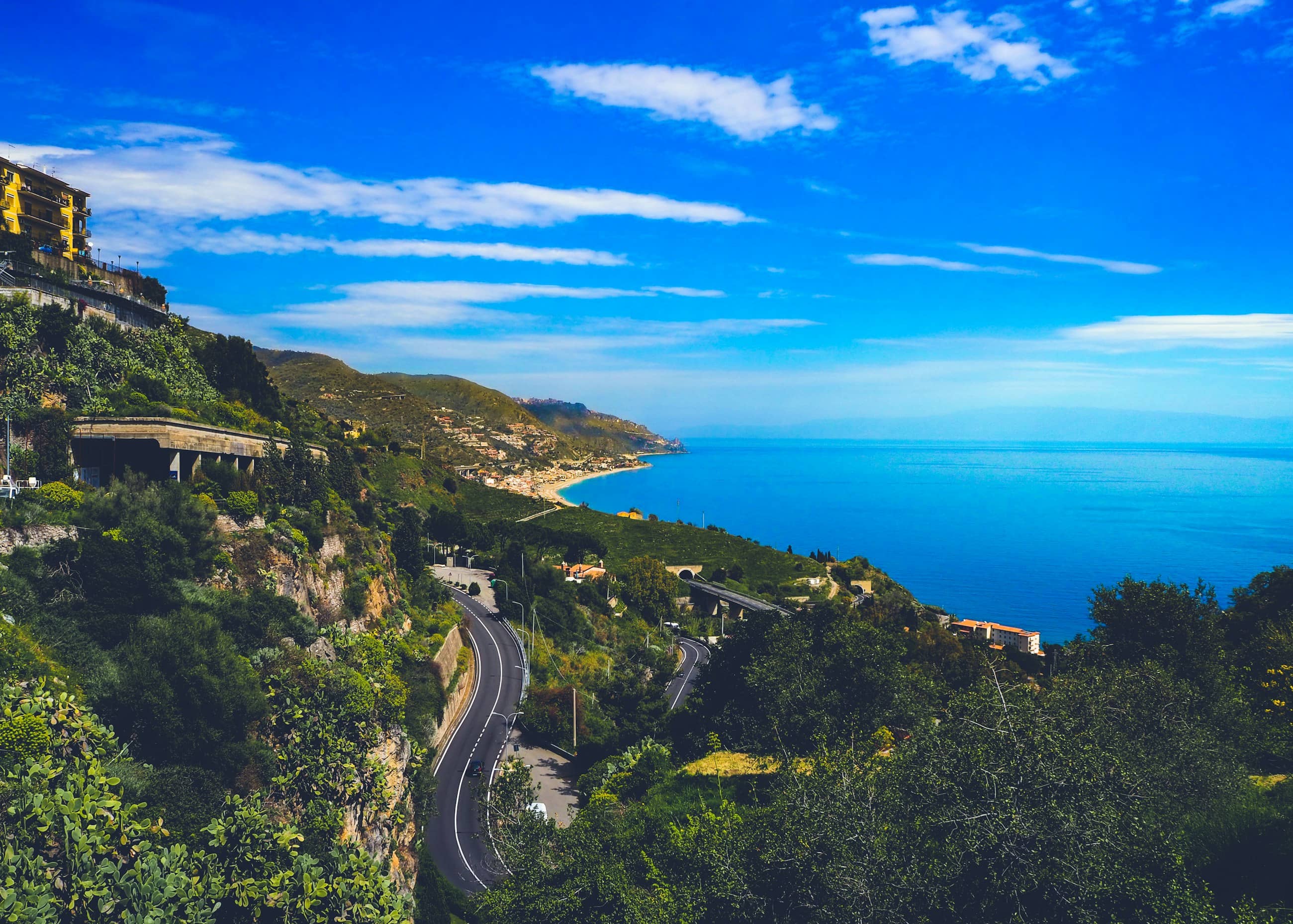 A road curving down through greenery to a long Sicilian coastline, open sea to the horizon.