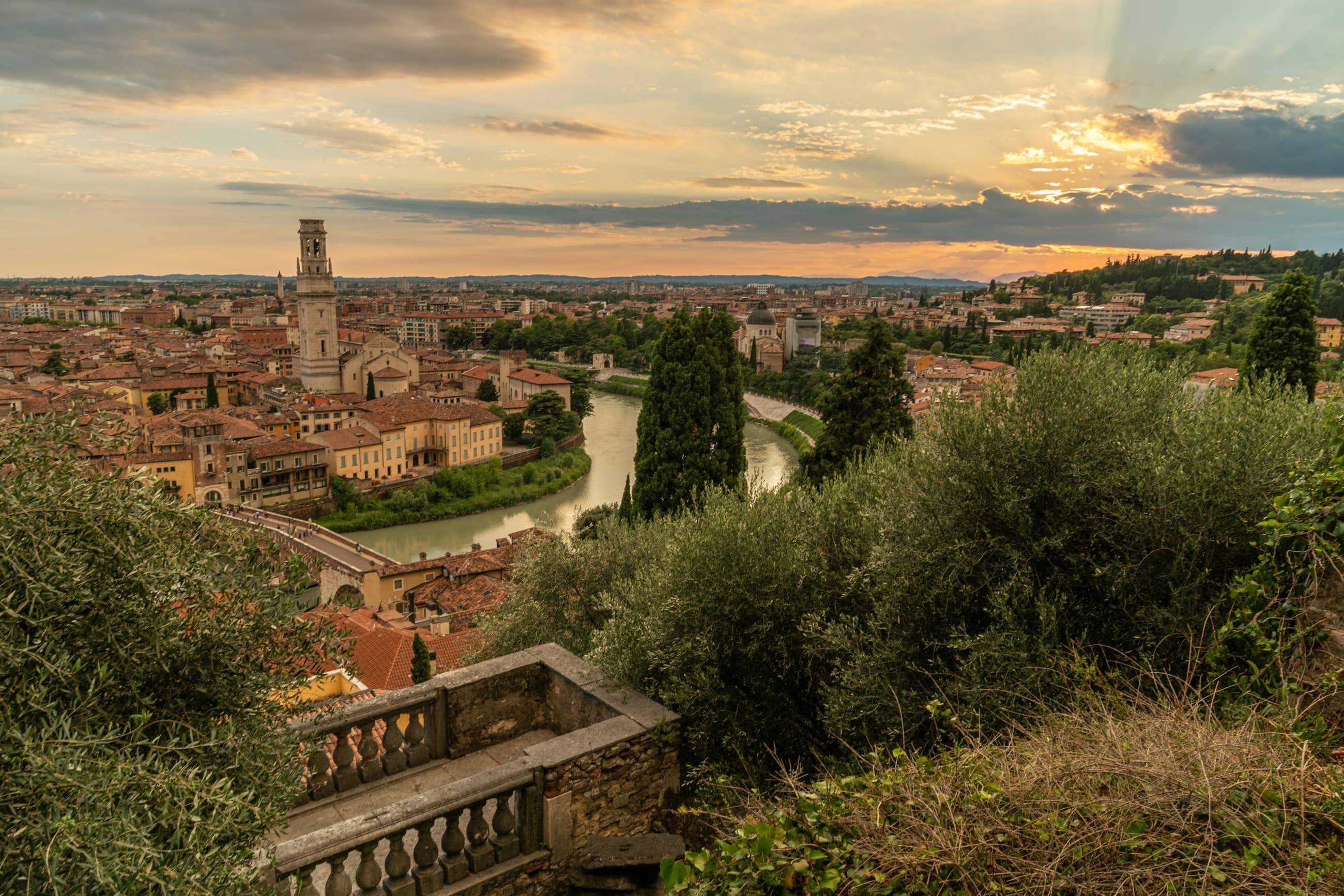 Verona at dusk from the hillside above it, the river curving through terracotta roofs and bell towers.